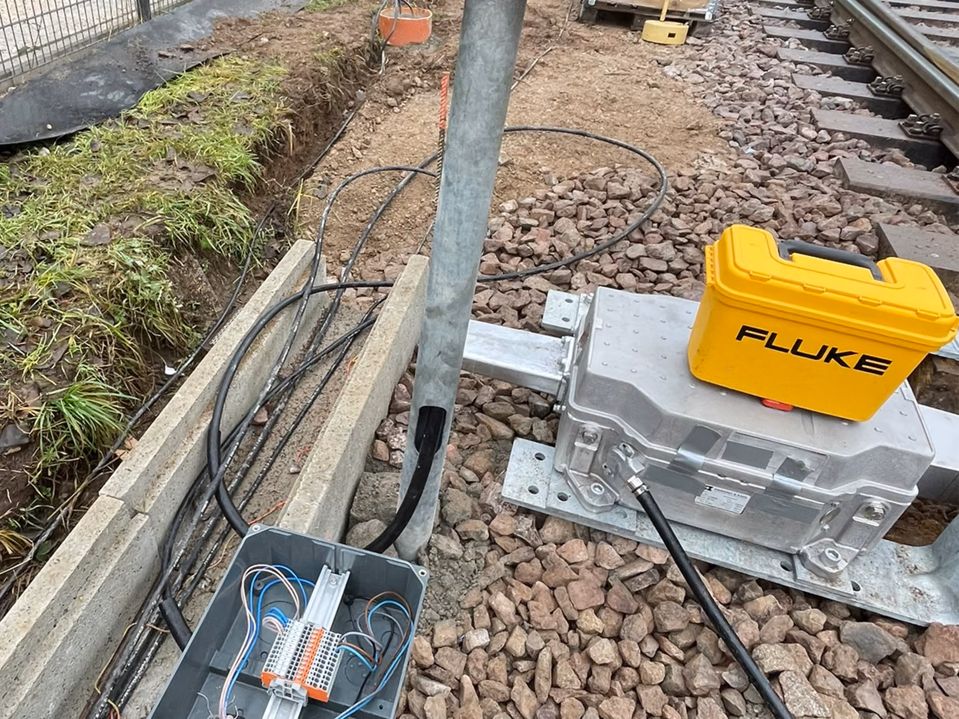 Open electrical junction box with visible wiring near railway tracks with ballast, accompanied by a metal electrical unit and a yellow Fluke carrying case on top.