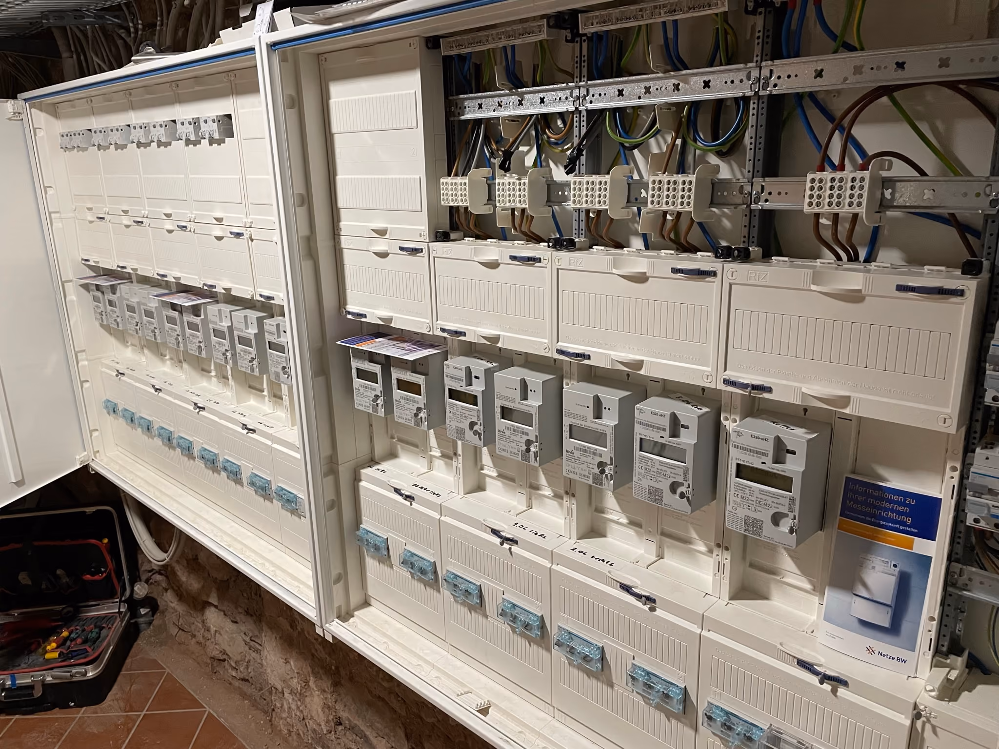 Electrical panel with multiple digital meters, wiring, and circuit breakers organized in rows inside a white metal enclosure mounted on a wall with a stone base.