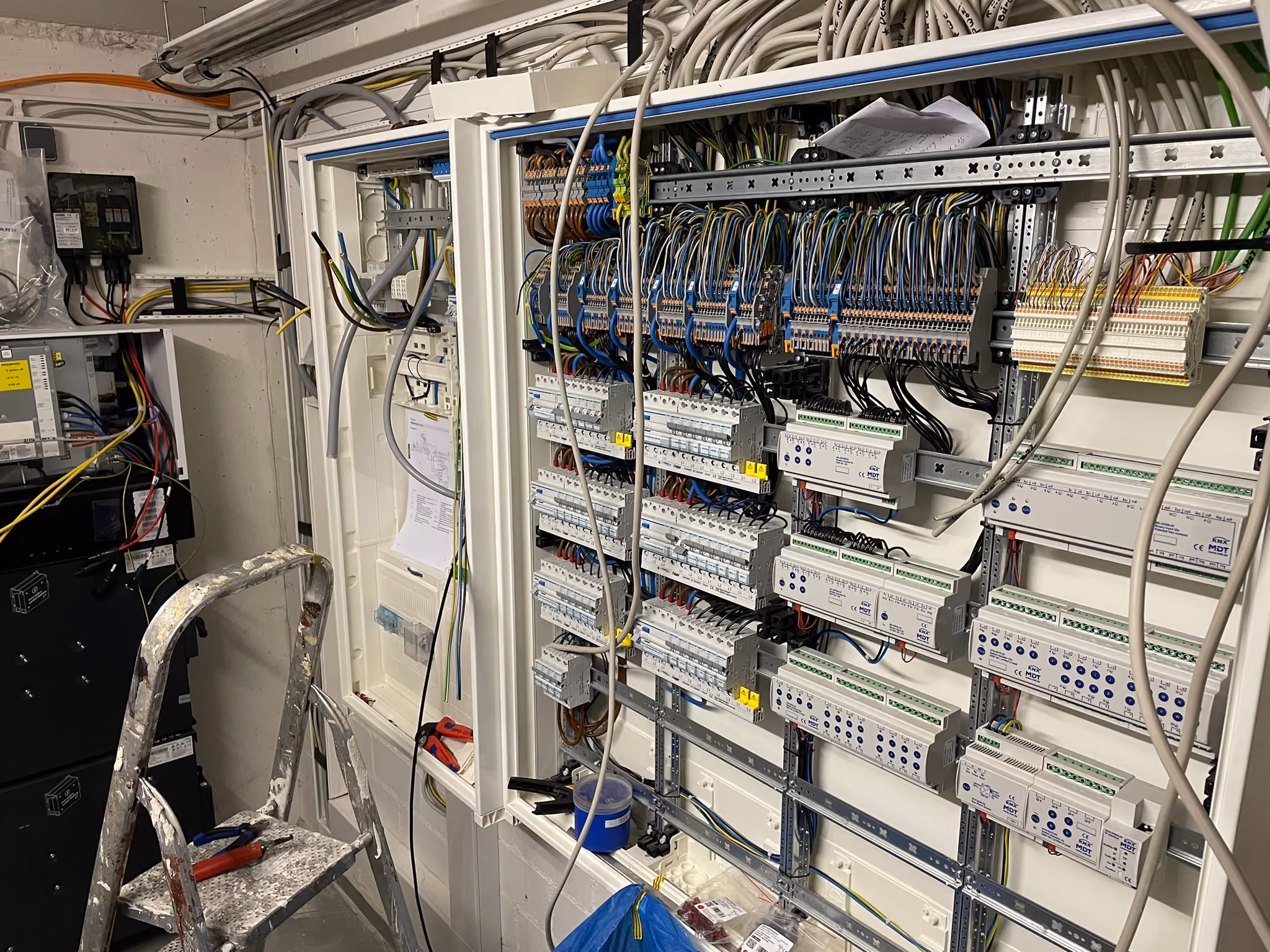 Electrical control panel with multiple circuit breakers, wiring, and terminal blocks mounted on a white wall, with cables hanging loosely and a ladder in front.