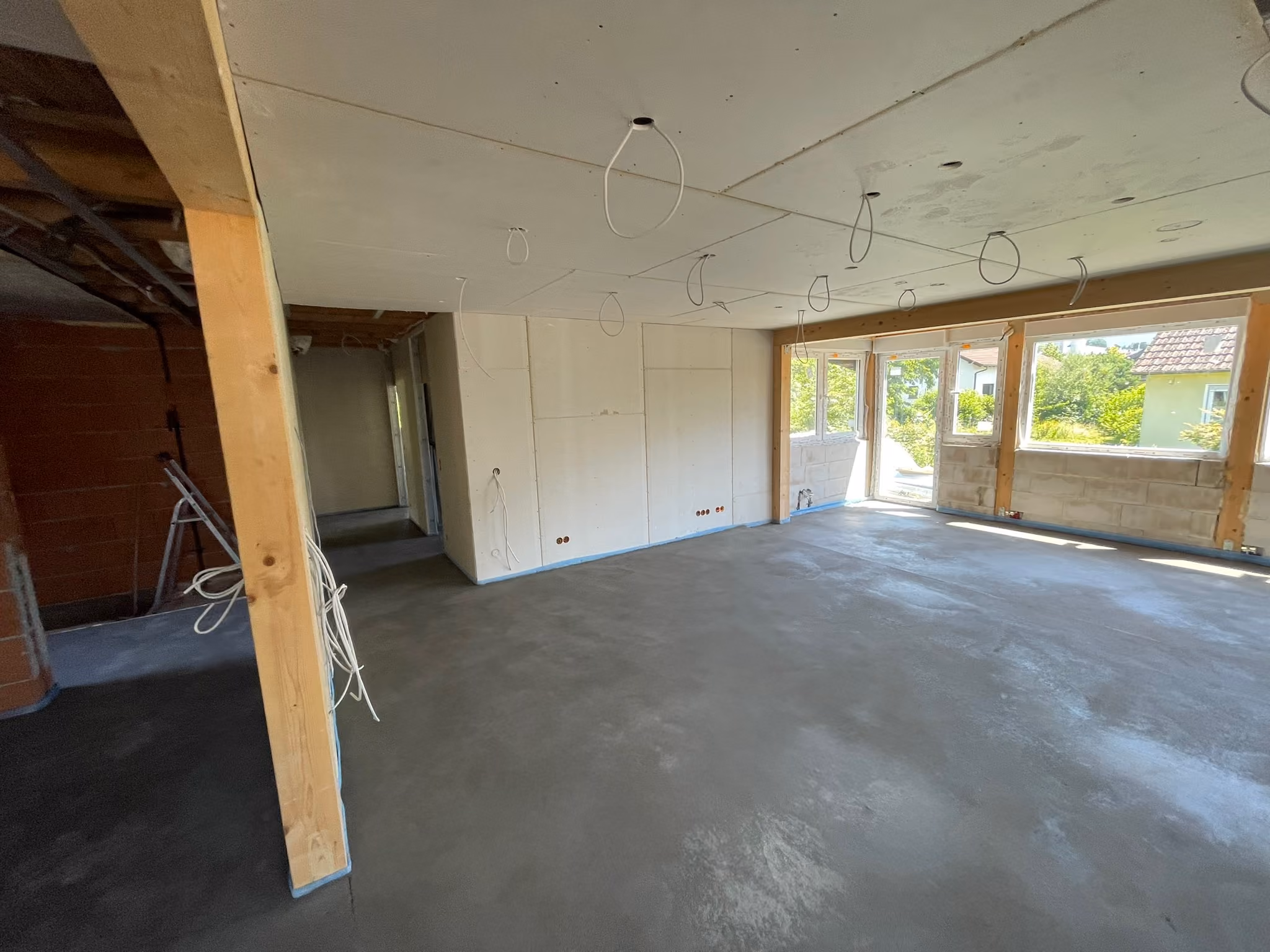 Interior of an unfinished room under construction with concrete floor, drywall partially installed, exposed wiring hanging from the ceiling, wooden support beams, and large open window spaces letting in daylight.