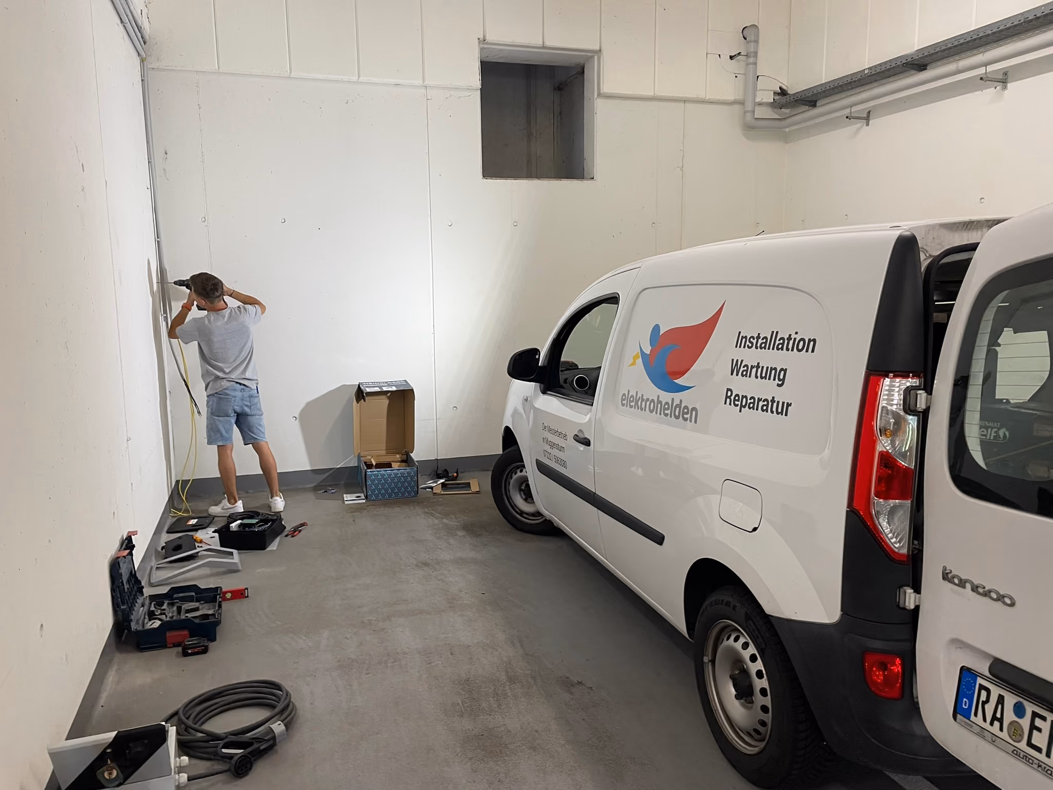 A man in a gray t-shirt and blue shorts is working on a white wall in a garage or workshop beside a white van with the logo and text 'elektrohelden' and phrases 'Installation Wartung Reparatur' on its side, surrounded by scattered tools and equipment on the concrete floor.